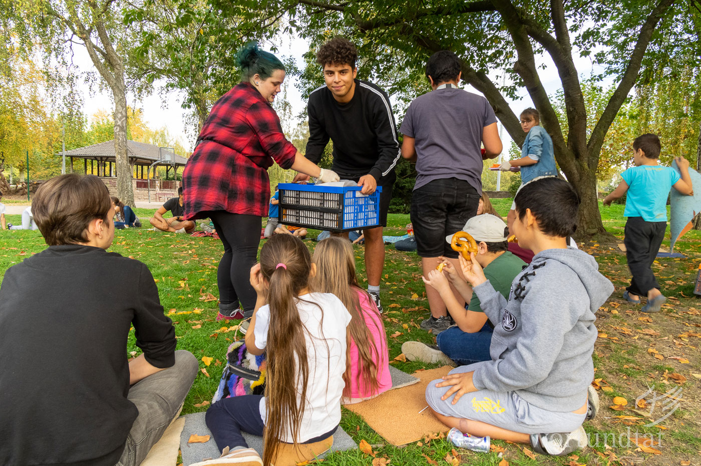 Bei allen Arbeiten fasst Achraf tatkräftig mit an. two young adults distributing fresh food to children that are sitting on  the meadow