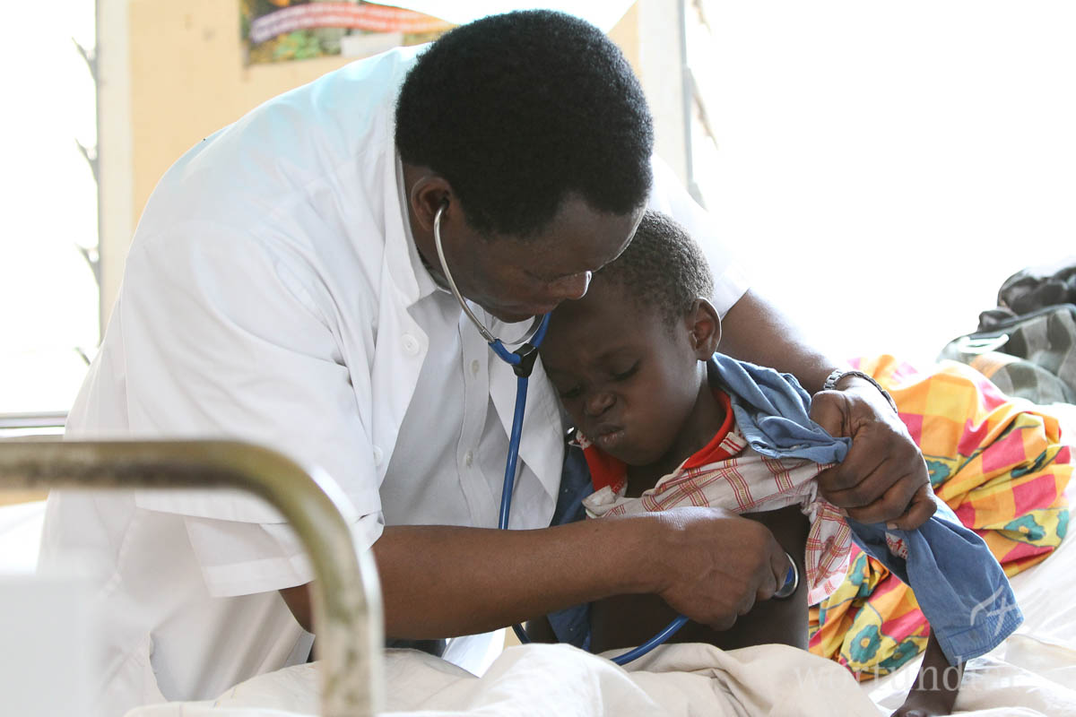 Seit das KIUMA-Hospital steht, haben Menschen in der Region Zugang zu guten medizinischen Leistungen. Arzt hält Jungen im Arm und horcht dabei die Lunge ab. Junge verzieht das Gesicht.