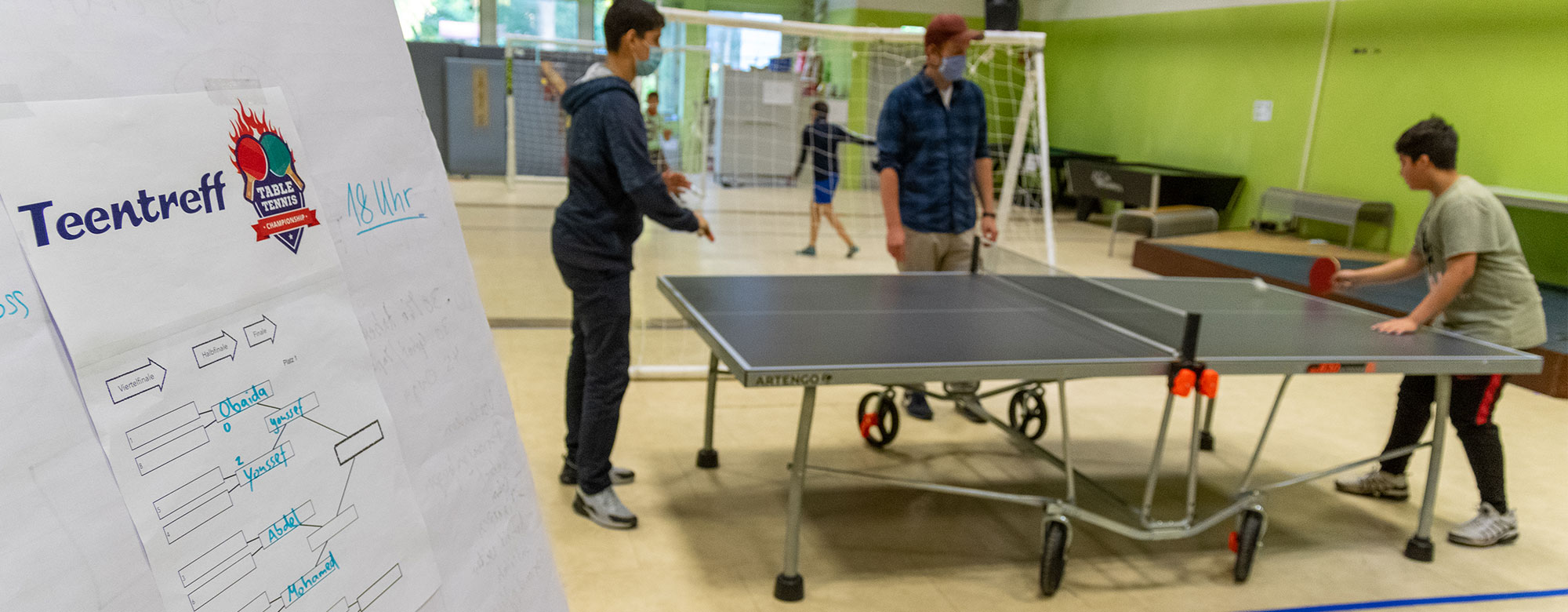 Im den großzügigen Räumen des Teentreff haben Jugendliche die Gelegenheit, sich auszutoben und Gemeinschaft zu erleben. Schild mit der Aufschrift Teentreff - im Hintergrund Tischtennnis spielende Jugendliche