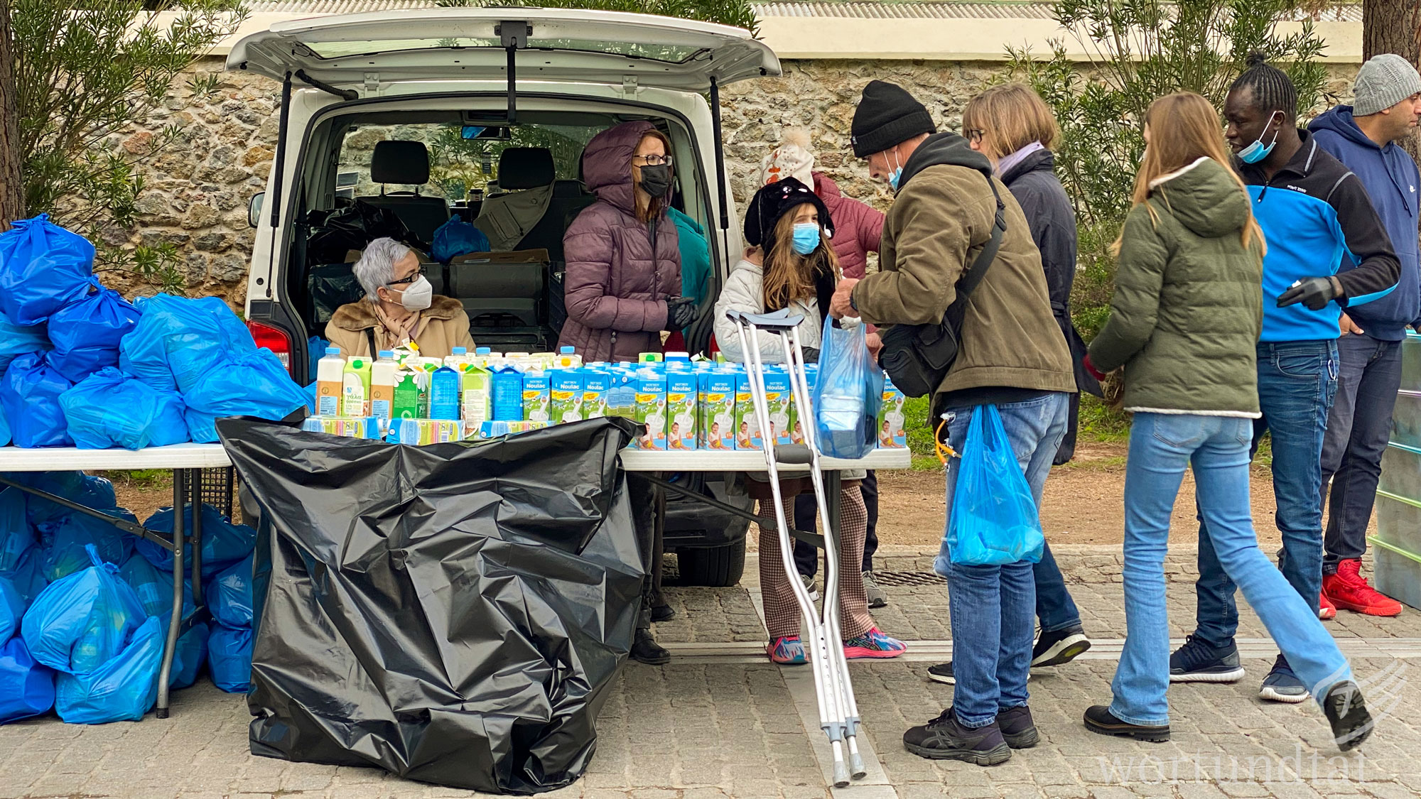 Helfer verteilen Mahlzeiten auf den Straßen von Athen an Menschen, die kein ausreichendes Einkommen haben: Obdachlose, Flüchtlinge, verarmte Rentner oder Menschen, die aus Osteuropa nach Athen gekommen sind. Ein Tisch mit Lebensmitteln, Menschen, die Schlange stehen