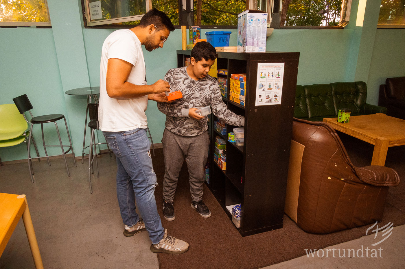 Gesellschaftsspiele werden von den Teens auch immer wieder gern genutzt. Viele kennen das von Zuhause kaum. Jugendlicher und Betreuer stehen an einem Regal mit Spielzeug