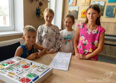 ein Junge sitzt mit Technikspielzeug am Tisch, drei Mädchen stehen daneben und schauen lächelnd in die Kamera