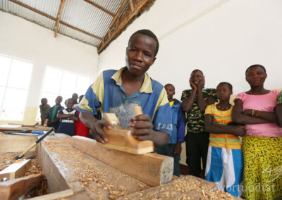 junger Mann hobelt ein Stück Holz - im Hintergrund Zuschauerinnen und Zuschauer