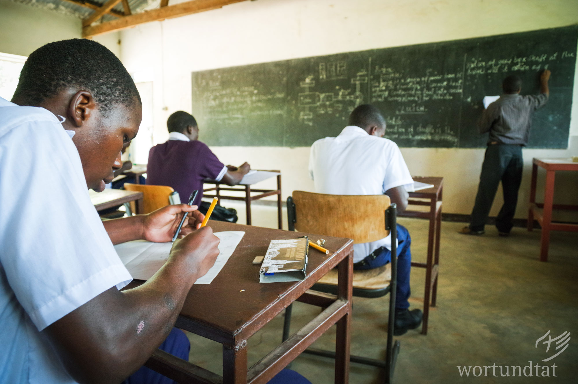 Young trainees receive technical drawing lessons in KIUMA. Young men sit at school desks and work