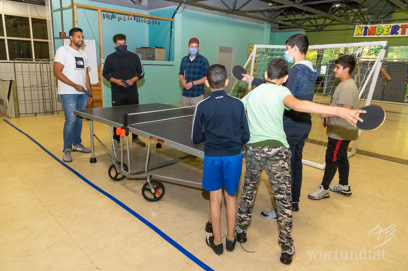 Bewegungsspiele sind gerade für Jugendliche eine gute Möglichkeit, zusammen aktiv zu sein. Gruppe von sieben jungen Männern beim Rundlauf an der Tischtennisplatte